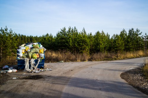 An overflowing blue wheelie bin situated on the edge of a gravel and dirt roadside area, filled with assorted waste materials including plastic bags, cardboard boxes, and miscellaneous refuse, some of which has spilled onto the ground beside the bin. The surrounding environment features a natural backdrop of dense green bushes and trees under a clear sky, with the bin positioned on a slight curve in the road that appears to be an unpaved rural or semi-rural pathway. The scene illustrates improper rubbish disposal, relevant to waste collection and clearance services in areas around Holborn and the broader London region, highlighting the need for professional rubbish removal such as that offered by Office Clearance Holborn.