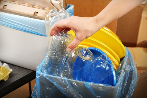 A close-up view of a person's hand gripping a clear plastic water bottle as part of a waste collection process. The bottle is situated inside a transparent plastic bag that contains various colourful household waste items, including yellow and blue plastic caps and containers. The background shows part of a room with wooden furniture and a dark surface, indicative of an indoor setting. The image highlights waste sorting or collection activities typical of rubbish removal services in the Holborn area, with a focus on recyclable plastic materials. The lighting is even, emphasizing the textures of the plastic bottles and caps, while the overall context suggests an organized rubbish collection or disposal task at a commercial or residential property in London.
