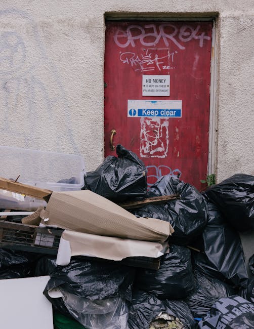 A large pile of black garbage bags, some partially torn or loosened, stacked against a building with a tiled exterior wall. The bags are filled with waste, and a few transparent plastic bottles are vi