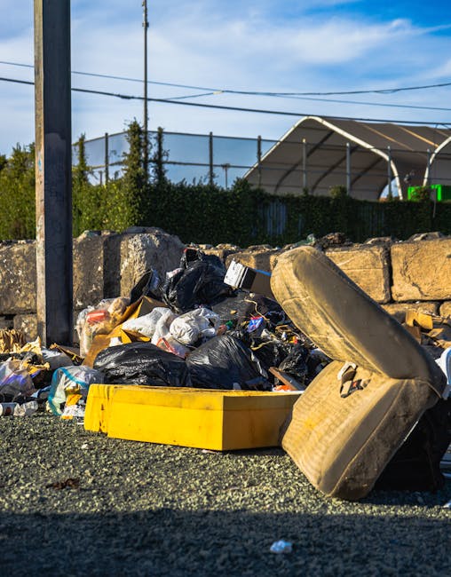 An aerial view of a large accumulation of scattered waste and debris located in a wooded area adjacent to a paved footpath and a raised hedge separating it from an adjacent road. The rubbish includes 