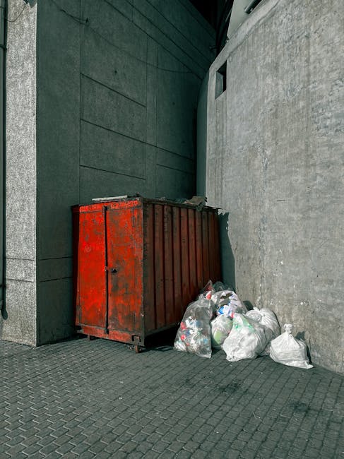 A black cylindrical trash bin positioned against a large, smooth beige stone wall in an indoor or underground setting. The top of the bin is overflowing with various rubbish items, including crumpled