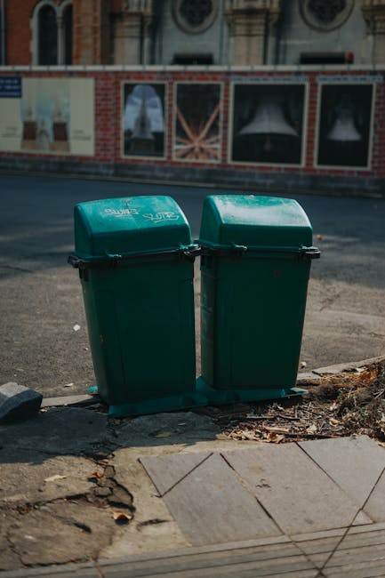 Two green plastic rubbish bins with closed lids are positioned side by side on a paved sidewalk curb, adjacent to a worn concrete area with visible dirt and small debris scattered at the base. The bin
